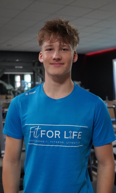 Junger Mann mit blauem Fit For Life Shirt laechelt leicht in einem Fitnessstudio